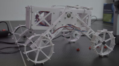 Biały model robota zbudowany z elementów wydrukowanych w technologii druku 3D stoi na stole. Konstrukcja ma lekką, ażurową obudowę i cztery duże koła z wypustkami, połączone z ramą za pomocą ruchomych ramion. Wewnątrz obudowy widoczne są przewody oraz elementy mechaniczne. Model prezentuje przykładowe zastosowanie druku 3D w budowie robotów i konstrukcji technicznych.