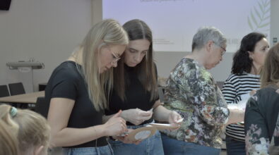 Uczestniczki warsztatów pracują przy stolikach, analizując materiały i wykonując zadanie praktyczne. Na pierwszym planie dwie kobiety koncentrują się na przygotowaniu elementów ćwiczenia, trzymając w dłoniach kartonowe podstawki i notatki. W tle widoczna jest prezentacja multimedialna z tytułem „Ułożenie jadłospisu na jeden dzień”, która wskazuje na edukacyjny charakter spotkania.