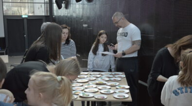 Uczestnicy warsztatów pracują w grupie przy stole, na którym rozłożone są materiały dydaktyczne w formie kart i ilustracji przedstawiających produkty spożywcze. Mężczyzna prowadzący zajęcia analizuje kartę z zadaniem i przekazuje wskazówki uczestnikom, wspierając ich w realizacji ćwiczenia. W działania zaangażowane są zarówno dzieci, jak i dorośli, co wskazuje na międzypokoleniowy charakter spotkania.