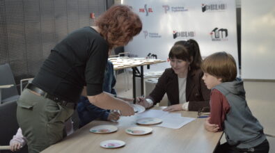 Uczestnicy warsztatów pracują przy stoliku, realizując zadanie polegające na układaniu jadłospisu na podstawie przygotowanych materiałów dydaktycznych. Prowadząca wspiera grupę, wskazując kolejne elementy ćwiczenia i udzielając wskazówek, natomiast uczestniczka zapisuje wypracowane rozwiązania. W działania aktywnie włącza się również dziecko, obserwując proces i współuczestnicząc w podejmowaniu decyzji.