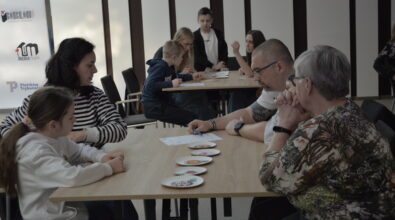 Uczestnicy warsztatów pracują w niewielkich grupach przy stolikach, wspólnie analizując zadanie i dobierając elementy jadłospisu na podstawie przygotowanych materiałów. Na pierwszym planie dorośli wraz z dzieckiem omawiają możliwe rozwiązania, podejmując decyzje dotyczące wyboru produktów. W tle widoczna jest kolejna grupa uczestników zaangażowanych w podobne ćwiczenie.