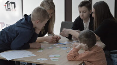 Grupa dzieci i dorosłych wspólnie pracuje przy stoliku, realizując zadanie warsztatowe polegające na dopasowywaniu elementów i układaniu spójnego schematu jadłospisu. Uczestnicy aktywnie dyskutują i analizują dostępne materiały, podejmując decyzje w oparciu o przedstawione informacje.