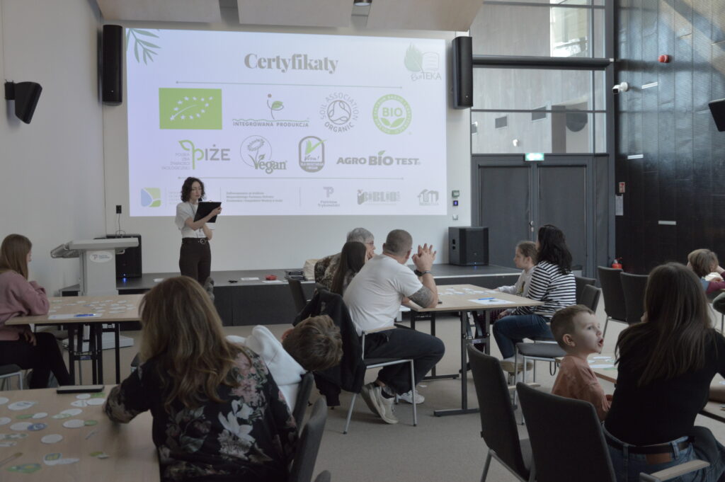 Prowadząca warsztaty omawia zagadnienia związane z certyfikatami jakości żywności, prezentując materiał na dużym ekranie multimedialnym. Na slajdzie widoczne są oznaczenia produktów ekologicznych i certyfikatów jakości, co wprowadza uczestników w temat świadomego wyboru żywności. Uczestnicy, pracujący przy stolikach w grupach międzypokoleniowych, aktywnie słuchają i jednocześnie realizują zadania praktyczne.