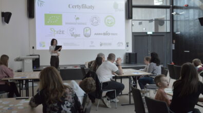 Prowadząca warsztaty omawia zagadnienia związane z certyfikatami jakości żywności, prezentując materiał na dużym ekranie multimedialnym. Na slajdzie widoczne są oznaczenia produktów ekologicznych i certyfikatów jakości, co wprowadza uczestników w temat świadomego wyboru żywności. Uczestnicy, pracujący przy stolikach w grupach międzypokoleniowych, aktywnie słuchają i jednocześnie realizują zadania praktyczne.