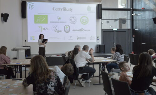 Prowadząca warsztaty omawia zagadnienia związane z certyfikatami jakości żywności, prezentując materiał na dużym ekranie multimedialnym. Na slajdzie widoczne są oznaczenia produktów ekologicznych i certyfikatów jakości, co wprowadza uczestników w temat świadomego wyboru żywności. Uczestnicy, pracujący przy stolikach w grupach międzypokoleniowych, aktywnie słuchają i jednocześnie realizują zadania praktyczne.