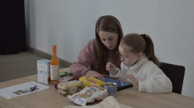 Kobieta wraz z dzieckiem pracują przy stoliku, analizując zadanie na tablecie edukacyjnym. Na blacie znajdują się różnorodne produkty spożywcze, które stanowią materiał pomocniczy do ćwiczenia z zakresu rozpoznawania składu i wartości żywności. Uczestniczki wspólnie omawiają wybory, podejmując decyzje dotyczące jakości produktów i ich miejsca w codziennym jadłospisie.