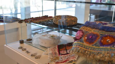 Przeszklona gablota ekspozycyjna zawiera ozdobne nakrycia głowy oraz elementy biżuterii. Na górnej półce leży haftowana czapka w złotych i czarnych kolorach oraz długi pas ozdobiony kolorowymi koralikami. Na dolnej półce znajduje się białe nakrycie głowy z tiulem i kryształkami, fragment haftowanej tkaniny oraz kilka par srebrnych kolczyków ułożonych w rzędzie. Po prawej stronie widoczne jest bogato zdobione, kolorowe nakrycie głowy z cekinami i złotą frędzlą. W tle przez duże okna wpada naturalne światło do wnętrza.