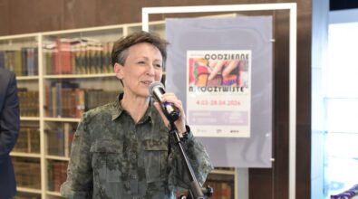 Kobieta z krótkimi ciemnymi włosami stoi przy statywie z mikrofonem i przemawia do zgromadzonych uczestników wydarzenia. Ma na sobie zieloną koszulę z delikatnym wzorem i uśmiecha się podczas wypowiedzi. Za nią widoczne są regały wypełnione książkami oraz stojak z plakatem informującym o wystawie „Codzienne nieoczywiste”, prezentowanej w terminie od 4 marca do 28 kwietnia 2024 roku. W lewej części kadru częściowo widoczna jest sylwetka innej osoby stojącej obok.