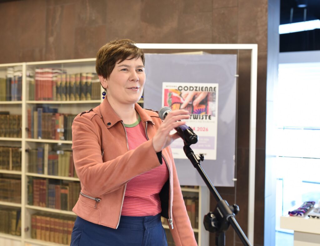 Kobieta z krótkimi włosami stoi przy statywie z mikrofonem i gestem dłoni poprawia jego ustawienie podczas przemówienia. Ubrana jest w jasnobrązową kurtkę, różowo-czerwoną bluzkę w paski oraz granatową spódnicę, a w uszach ma długie kolczyki. W tle widoczne są regały z książkami oraz plansza z plakatem wystawy „Codzienne nieoczywiste” z informacją o terminie ekspozycji od 4 marca do 28 kwietnia 2024 roku. Po prawej stronie znajduje się podświetlona gablota ekspozycyjna