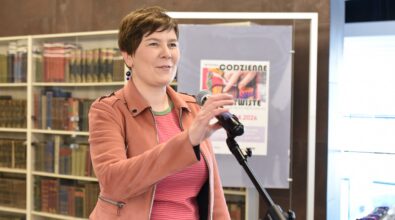 Kobieta z krótkimi włosami stoi przy statywie z mikrofonem i gestem dłoni poprawia jego ustawienie podczas przemówienia. Ubrana jest w jasnobrązową kurtkę, różowo-czerwoną bluzkę w paski oraz granatową spódnicę, a w uszach ma długie kolczyki. W tle widoczne są regały z książkami oraz plansza z plakatem wystawy „Codzienne nieoczywiste” z informacją o terminie ekspozycji od 4 marca do 28 kwietnia 2024 roku. Po prawej stronie znajduje się podświetlona gablota ekspozycyjna