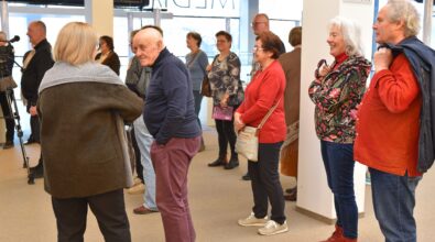 Grupa dorosłych osób stoi w przestronnym holu i rozmawia podczas wydarzenia. Kilka osób prowadzi rozmowę w małych grupach, inni stoją i obserwują to, co dzieje się w sali. Wśród uczestników widać m.in. starszego mężczyznę w granatowej bluzie i bordowych spodniach oraz kobietę w czerwonym swetrze z torebką przewieszoną przez ramię. Po prawej stronie stoją dwie osoby – kobieta w kwiatowej kurtce i czerwonych butach oraz mężczyzna w czerwonej koszuli z zarzuconą na ramię kurtką. W tle znajdują się duże okna, filary budynku oraz znak informujący o toaletach. W lewej części kadru widoczna jest kamera ustawiona na statywie.