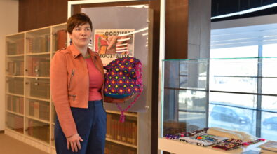 Kobieta stoi obok przeszklonej gabloty ekspozycyjnej i trzyma w rękach kolorowy, haftowany worek z geometrycznym wzorem i ozdobnymi sznurkami. Ubrana jest w jasnobrązową kurtkę oraz różową bluzkę w paski i zwraca się do uczestników wydarzenia, prezentując przedmiot. Za nią widoczna jest plansza z plakatem wystawy „Codzienne nieoczywiste”, a w tle znajdują się regały z książkami. Na blacie gabloty obok leżą haftowane tkaniny oraz inne drobne elementy ekspozycji.