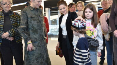 Chłopiec stoi wśród uczestników wydarzenia i ma na głowie haftowaną czapkę ozdobioną kolorowymi wzorami. Obok niego stoi kobieta w czarnej marynarce, która poprawia nakrycie głowy na jego głowie. Wokół zgromadzili się dorośli oraz dzieci obserwujący sytuację z zainteresowaniem i uśmiechem. Po lewej stronie widoczna jest kobieta w długiej zielonej sukni w roślinny wzór, a obok niej starsza kobieta w okularach i w kratkowanym płaszczu. W tle stoją kolejne osoby uczestniczące w wydarzeniu.