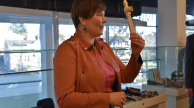 Kobieta z krótkimi włosami stoi obok przeszklonej gabloty ekspozycyjnej i trzyma w dłoni jasny, drewniany instrument przypominający flet prosty. Ubrana jest w jasnobrązową kurtkę oraz różową bluzkę w paski i prezentuje przedmiot podczas rozmowy z uczestnikami wydarzenia. W tle widoczne są duże okna oraz gablota z różnymi eksponatami, wśród których znajdują się ozdobne nakrycia głowy i drobne przedmioty dekoracyjne.