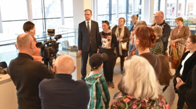 Grupa kilkunastu osób stoi wokół kobiety przemawiającej podczas wydarzenia. Uczestnicy ustawieni są półkolem i słuchają wypowiedzi, a część z nich obserwuje nagranie realizowane przez operatora kamery stojącego z przodu. Wśród zgromadzonych znajduje się mężczyzna w garniturze, kobieta w czarnej koszulce z graficznym wizerunkiem twarzy oraz kilka osób w płaszczach i swetrach. W centrum stoi również chłopiec w czapce z daszkiem i koszuli w zieloną kratę. W tle widoczne są duże okna, przeszklone gabloty z eksponatami oraz nowoczesne oświetlenie sufitowe.