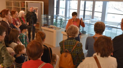 Kobieta prowadząca spotkanie stoi przy przeszklonych gablotach i prezentuje uczestnikom element stroju lub ozdobę trzymaną w dłoni. Wokół niej zgromadziła się grupa osób – dorośli oraz dzieci – ustawieni półkolem i słuchający jej wypowiedzi. Z lewej strony operator kamery nagrywa wydarzenie, a obok stoją uczestnicy obserwujący prezentację. W gablotach znajdują się kolorowe tkaniny i elementy strojów, a w tle widać duże okna oraz regały z książkami.