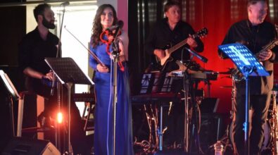 Zespół występuje na scenie z wokalistami i instrumentalistami. Wokalistka w niebieskiej sukience stoi na środku, muzycy Jasne oświetlenie sceniczne.