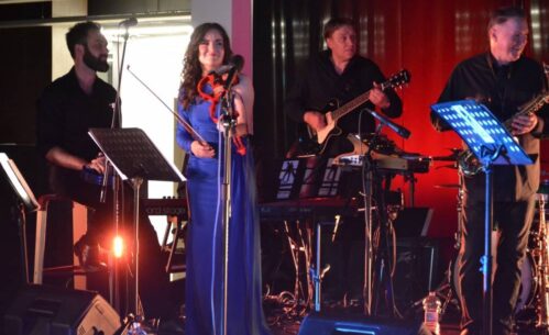 Zespół występuje na scenie z wokalistami i instrumentalistami. Wokalistka w niebieskiej sukience stoi na środku, muzycy Jasne oświetlenie sceniczne.