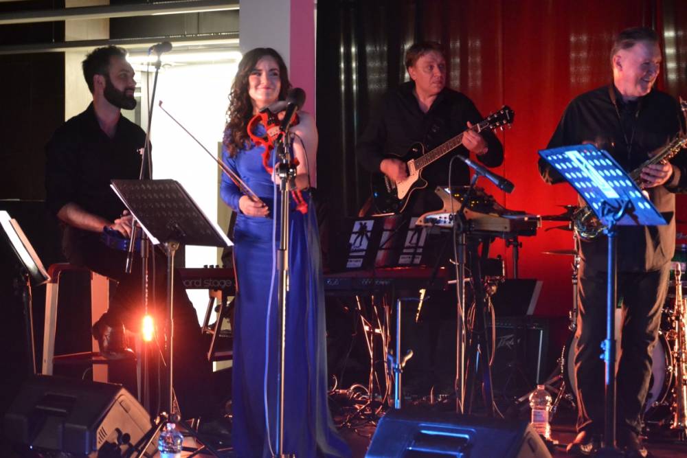 Zespół występuje na scenie z wokalistami i instrumentalistami. Wokalistka w niebieskiej sukience stoi na środku, muzycy Jasne oświetlenie sceniczne.