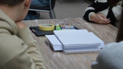 Stół przykryty folią ochronną, na którym leżą arkusze białego papieru, taśma malarska, czarny klips na dokumenty, klej w sztyfcie i opakowanie kolorowych flamastrów. Przy stole siedzą osoby, których twarze nie są widoczne, ubrane w jasne i ciemne ubrania