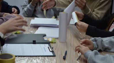 Stół pokryty folią ochronną, na którym znajdują się arkusze papieru w kratkę, taśma malarska, klej w tubce, opakowanie kolorowych flamastrów oraz czarna teczka z dokumentami i długopisem. Kilka osób siedzi przy stole, a ich dłonie są zajęte składaniem i układaniem papieru. Widoczne różne kolory ubrań uczestników