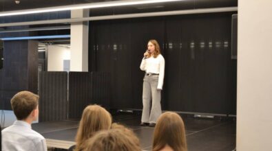 Dziewczyna w białej bluzce stoi na scenie, trzymając mikrofon w dłoni. Za nią znajduje się czarna kotara, a z przodu, na widowni, siedzi publiczność. W tle widoczne są dwa głośniki.