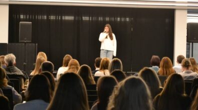 Dziewczyna stoi na scenie, trzymając mikrofon w dłoni. Za nią znajduje się czarna kotara, a z przodu, na widowni, siedzi publiczność. W tle widoczne są dwa głośniki.