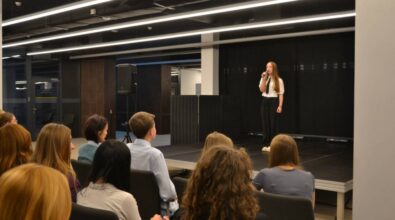 Dziewczyna w białej koszuli z czarnym krawatem stoi na scenie, trzymając mikrofon w dłoni. Za nią znajduje się czarna kotara, a z przodu, na widowni, siedzi publiczność. W tle widoczne są dwa głośniki.