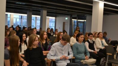 Publiczność siedząca w jasnej przestrzeni. W pierwszych rzędach widoczni są młodzi ludzie oraz osoby dorosłe, w tle zaś widać okna z widokiem na budynki miasta. W sali znajdują się białe filary i ciemny sufit z elementami technicznymi.