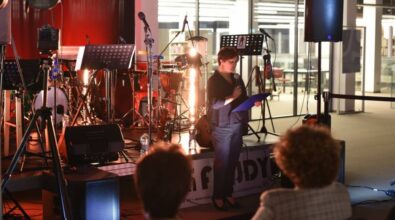 Osoba przemawiająca z mikrofonem, w tle scena z instrumentami muzycznymi. Z przodu widać ludzi siedzących na krzesłach.