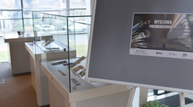 Sala ze szklanymi gablotami zawierającymi zakładki. Duży szyld na pierwszym planie przedstawia plakat promujący wystawę. Okno w tle wpuszcza naturalne światło.