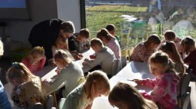 Grupa małych dzieci siedzących przy stołach, zaangażowanych w rysowanie. Kobieta pochyla się, aby pomóc jednemu dziecku. Przez duże okno wpada światło słoneczne.