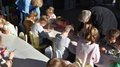 Dzieci siedzą przy niskich stołach, skupione na rysowaniu. Dwie kobiety stoją w pobliżu, pochylając się i pomagając im. Sala jest dobrze oświetlona, ​​panuje w niej atmosfera zabawy i edukacji.