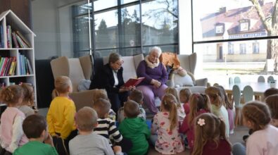 Dwie kobiety siedzą w fotelach i czytają książkę na głos grupie małych dzieci siedzących na podłodze w bibliotece. Pokój jest jasny, z dużymi oknami, a dzieci uważnie słuchają, otoczone regałami z książkami i pluszowymi zabawkami.
