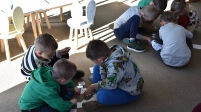 Dzieci siedzą na podłodze w parach, pracując nad zadaniami na karteczkach. Małe krzesła i stoły znajdują się w tle. Pomieszczenie jest dobrze oświetlone światłem naturalnym.