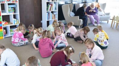 Dzieci siedzą na podłodze biblioteki, bawią się i są zaangażowane w różne aktywności. W pobliżu znajdują się półki z książkami i zabawkami. W tle dwie starsze osoby siedzą w fotelach i czytają. Przestrzeń jest jasna i zachęcająca.