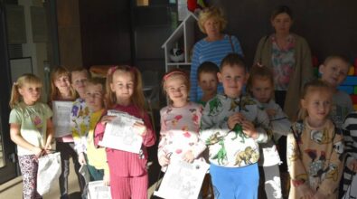 Grupa małych dzieci, niektóre trzymające rysunki, stoi razem z dwoma dorosłymi w pokoju. Dzieci mają na sobie kolorowe stroje, a w pokoju są zabawki i materiały do aktywności. Na półce w tle widać pluszową zabawkę.