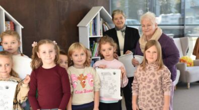 Grupa dzieci trzymających rysunki stoi w dobrze oświetlonym pokoju z regałami na książki w tle. Dwie starsze kobiety uśmiechają się za nimi, a duże okna pozwalają na wypełnienie przestrzeni naturalnym światłem.