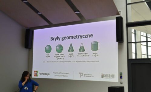 Prezentacja na temat brył geometrycznych wyświetlana na dużym ekranie. Zawiera ilustracje i wzory dotyczące kuli, odcinka kulistego, stożka, stożka ściętego oraz walca. Na dole ekranu logotypy organizatorów i sponsorów: Fundacja mBanku, Piotrków Trybunalski oraz Biblioteka w Piotrkowie Trybunalskim. Po lewej stronie kobieta w niebieskiej bluzce trzyma mikrofon, stojąc obok ekranu w nowoczesnym wnętrzu z przeszklonymi ścianami