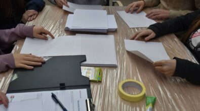 Stół pokryty folią ochronną, na którym znajdują się arkusze papieru w kratkę, taśma malarska, klej w tubce, opakowanie kolorowych flamastrów oraz czarna teczka z dokumentami i długopisem. Kilka osób siedzi przy stole, a ich dłonie są zajęte składaniem i układaniem papieru. Widoczne różne kolory ubrań uczestników