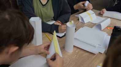 Stół pokryty folią ochronną, na którym znajdują się arkusze papieru w kratkę, taśma malarska, klej w tubce, opakowanie kolorowych flamastrów oraz czarna teczka z dokumentami i długopisem. Kilka osób siedzi przy stole, a ich dłonie są zajęte składaniem i układaniem papieru. Widoczne różne kolory ubrań uczestników