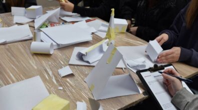 Stół przykryty folią ochronną, na którym leżą arkusze białego papieru, taśma malarska, czarny klips na dokumenty, klej w sztyfcie i opakowanie kolorowych flamastrów. Przy stole siedzą osoby, których twarze nie są widoczne, ubrane w jasne i ciemne ubrania