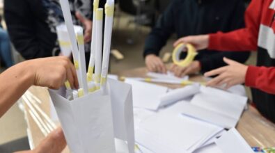 Stół, na którym znajdują się arkusze białego papieru, zwinięte w rulony i połączone taśmą malarską. Kilka osób pracuje nad tworzeniem konstrukcji z papieru, używając taśmy malarskiej. Widoczne są ręce uczestników, jedna osoba trzyma taśmę, a inna wkłada rulony do papierowego pojemnika. W tle rozmazane sylwetki osób i krzesła.