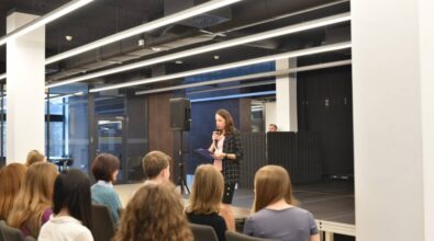 Kobieta w marynarce w kratę i różowej bluzce stoi na scenie, trzymając mikrofon i kartki, prowadząc wydarzenie lub przemówienie. W tle widać nowoczesne wnętrze z industrialnym sufitem i przeszklonymi ścianami. Na pierwszym planie siedzi grupa słuchaczy, skupionych na wystąpieniu. Scena jest wyposażona w głośnik, a w tle widoczny jest mężczyzna stojący za przegrodą akustyczną.