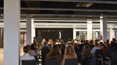 Widok na salę konferencyjną z perspektywy tylnych rzędów. Na scenie stoi młody mężczyzna z mikrofonem, za nim znajduje się czarna kotara. Publiczność siedzi na krzesłach, a po bokach widoczne są białe filary oraz regały z książkami. Sufit jest ciemny z widocznymi elementami oświetlenia i wentylacji.