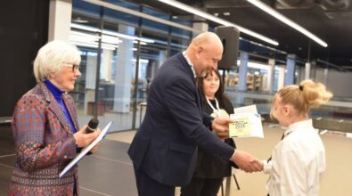 Osoby na scenie w trakcie uroczystości. Starsza kobieta z białymi włosami w kolorowej marynarce trzyma mikrofon i kartkę, mężczyzna w garniturze wręcza dyplom młodej dziewczynce ubranej w białą koszulę. W tle kobieta w ciemnym stroju oraz nowoczesne wnętrze z przeszklonymi ścianami i oświetleniem