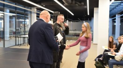 Na zdjęciu widzimy moment wręczania nagrody lub dyplomu w przestrzeni nowoczesnego wnętrza biblioteki. Mężczyzna w garniturze podaje rękę młodej dziewczynie z długimi blond włosami, ubranej w fioletowy sweter i jasnoszarą sukienkę. W tle mężczyzna z brodą w skórzanej kurtce trzyma mikrofon i przemawia. Po prawej stronie widać kilka siedzących osób, które obserwują wydarzenie. Wnętrze jest jasne, z nowoczesnym oświetleniem i przeszklonymi elementami.