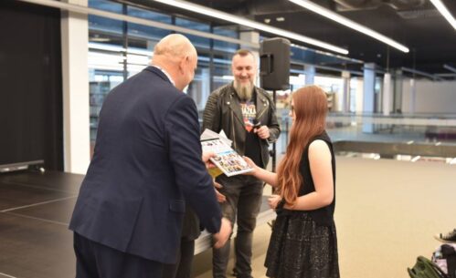 Na zdjęciu widać uroczystość wręczania nagród w przestrzeni nowoczesnego wnętrza, w bibliotece. Po lewej stronie mężczyzna w garniturze przekazuje dyplom dziewczynie z długimi rudymi włosami, ubranej w czarną sukienkę i kamizelkę. W tle mężczyzna z brodą i skórzaną kurtką trzyma mikrofon i uśmiecha się, obserwując sytuację. Widać również scenę oraz nowoczesne oświetlenie i przeszklone elementy wnętrza.
