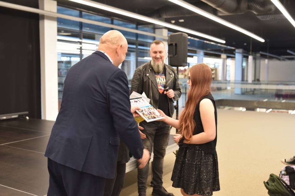 Na zdjęciu widać uroczystość wręczania nagród w przestrzeni nowoczesnego wnętrza, w bibliotece. Po lewej stronie mężczyzna w garniturze przekazuje dyplom dziewczynie z długimi rudymi włosami, ubranej w czarną sukienkę i kamizelkę. W tle mężczyzna z brodą i skórzaną kurtką trzyma mikrofon i uśmiecha się, obserwując sytuację. Widać również scenę oraz nowoczesne oświetlenie i przeszklone elementy wnętrza.