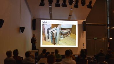 Na zdjęciu znajduje się prelegent stojący na scenie z mikrofonem, prezentujący slajd z wizualizacją książek ustawionych na półce. Sala jest wypełniona widzami, którzy siedzą na krzesłach i oglądają prezentację na dużym ekranie. W tle widać nowoczesne oświetlenie sceniczne i minimalistyczny wystrój wnętrza.